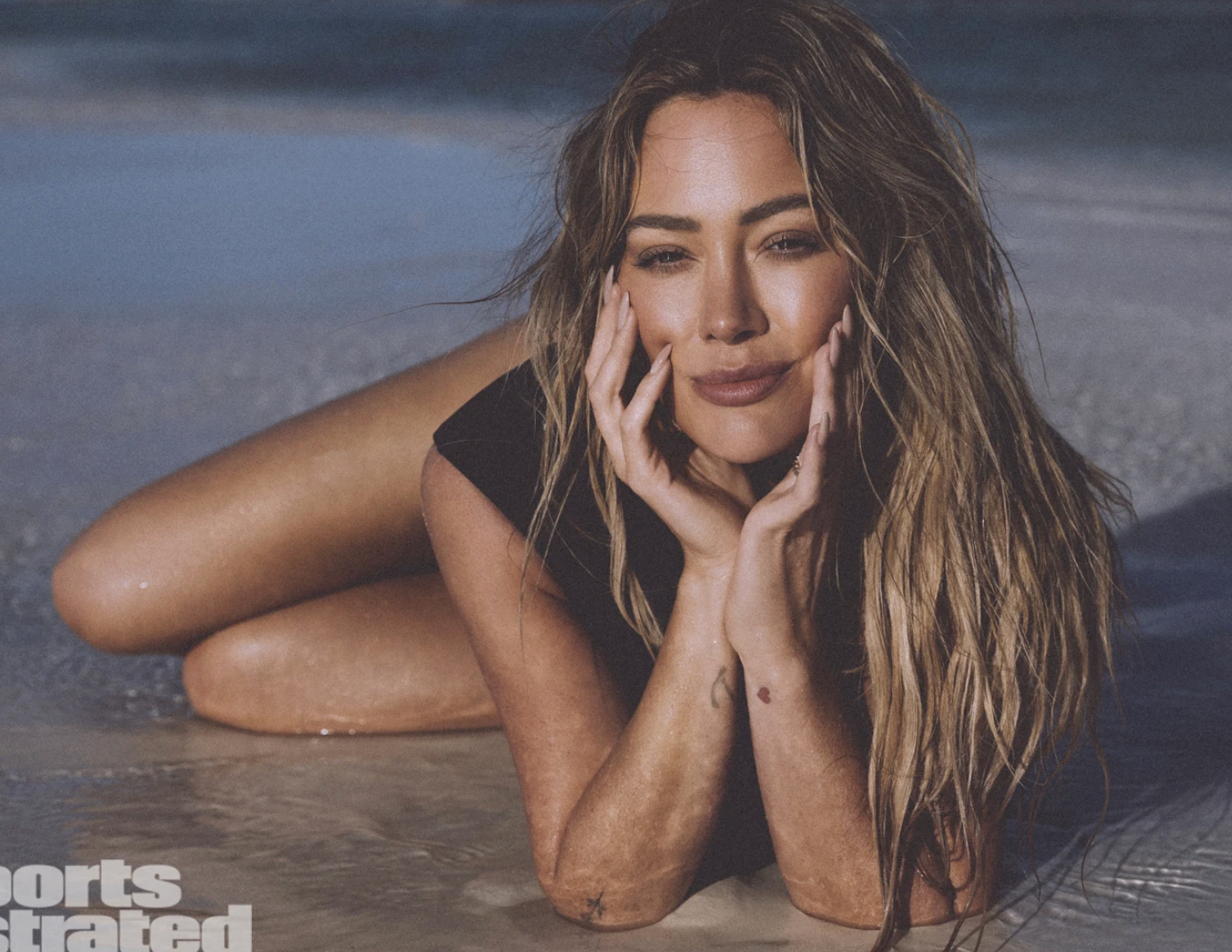 Woman with long wavy hair lying in shallow surf, hands cupping her face, wearing a black swimsuit on a sunny beach.