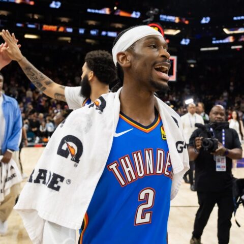 Basketball player in a blue Thunder jersey with towel over shoulders greets teammates on a celebratory court while photographers film nearby.