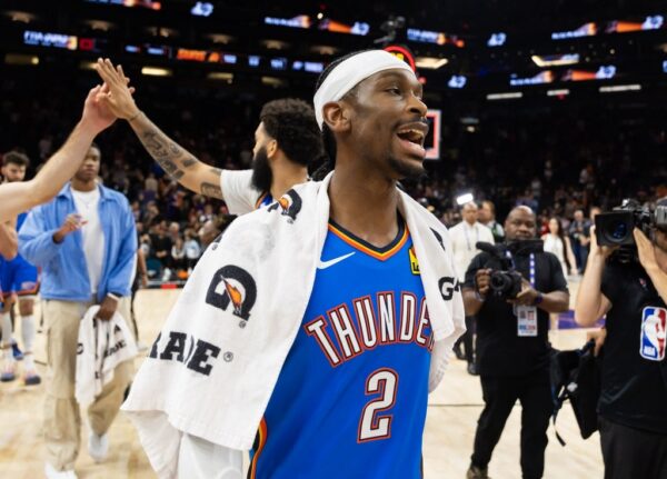 Basketball player in a blue Thunder jersey with towel over shoulders greets teammates on a celebratory court while photographers film nearby.