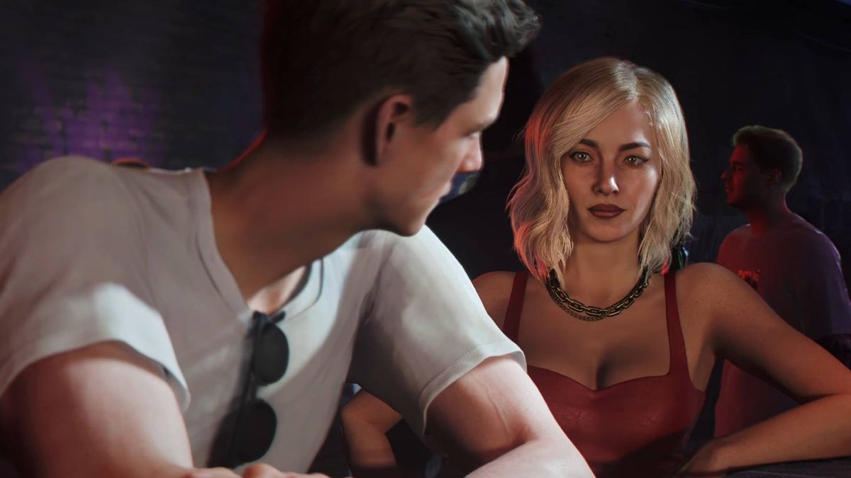 Man in a white shirt chats with a blonde woman in a red top at a dim bar setting, both looking at each other attentively.