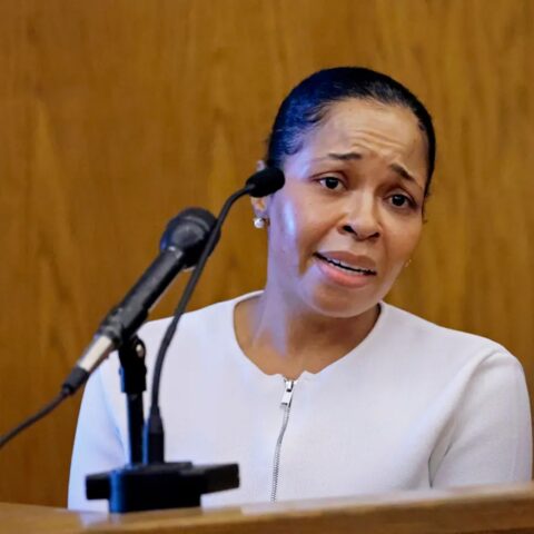 Woman in a white top standing at a wooden lectern, speaking into a microphone with a concerned expression.