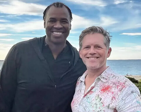 Two men smiling for a selfie on a beach, one in a black shirt and the other in a light patterned shirt, ocean and blue sky behind them.
