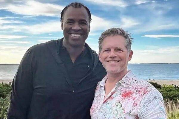 Two men smiling for a selfie on a beach, one in a black shirt and the other in a light patterned shirt, ocean and blue sky behind them.