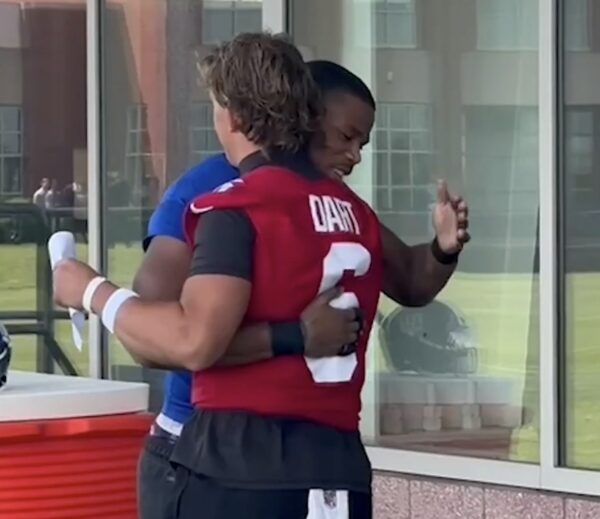 Two football players share a celebratory hug, one in a red jersey with the number 2 and name on the back, the other in blue, standing outside glass doors.