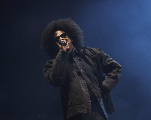 Male singer wearing sunglasses, microphone in hand, performing on a blue-lit stage.