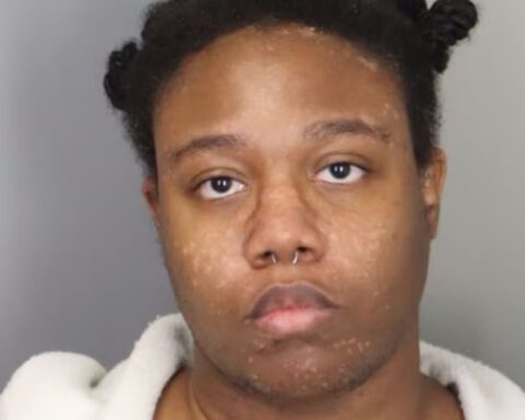 Mugshot-style portrait of a young person facing the camera, wearing a light robe, with a septum piercing.