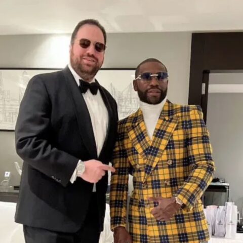 Two men in formal attire posing together at an event; one in a black tuxedo and bow tie, the other in a yellow plaid suit and white turtleneck, both wearing sunglasses.