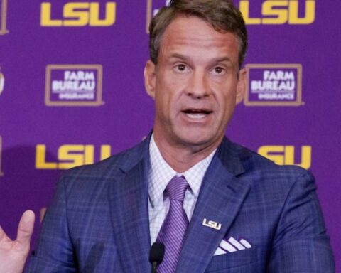 Man in a blue checked suit and purple tie speaks at a podium with an LSU backdrop behind him.