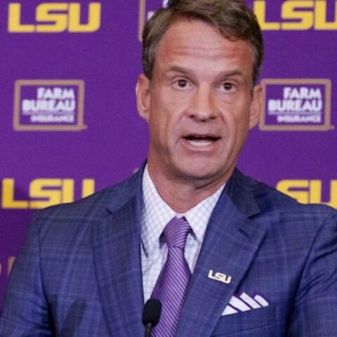 Man in a blue checked suit and purple tie speaks at a podium with an LSU backdrop behind him.