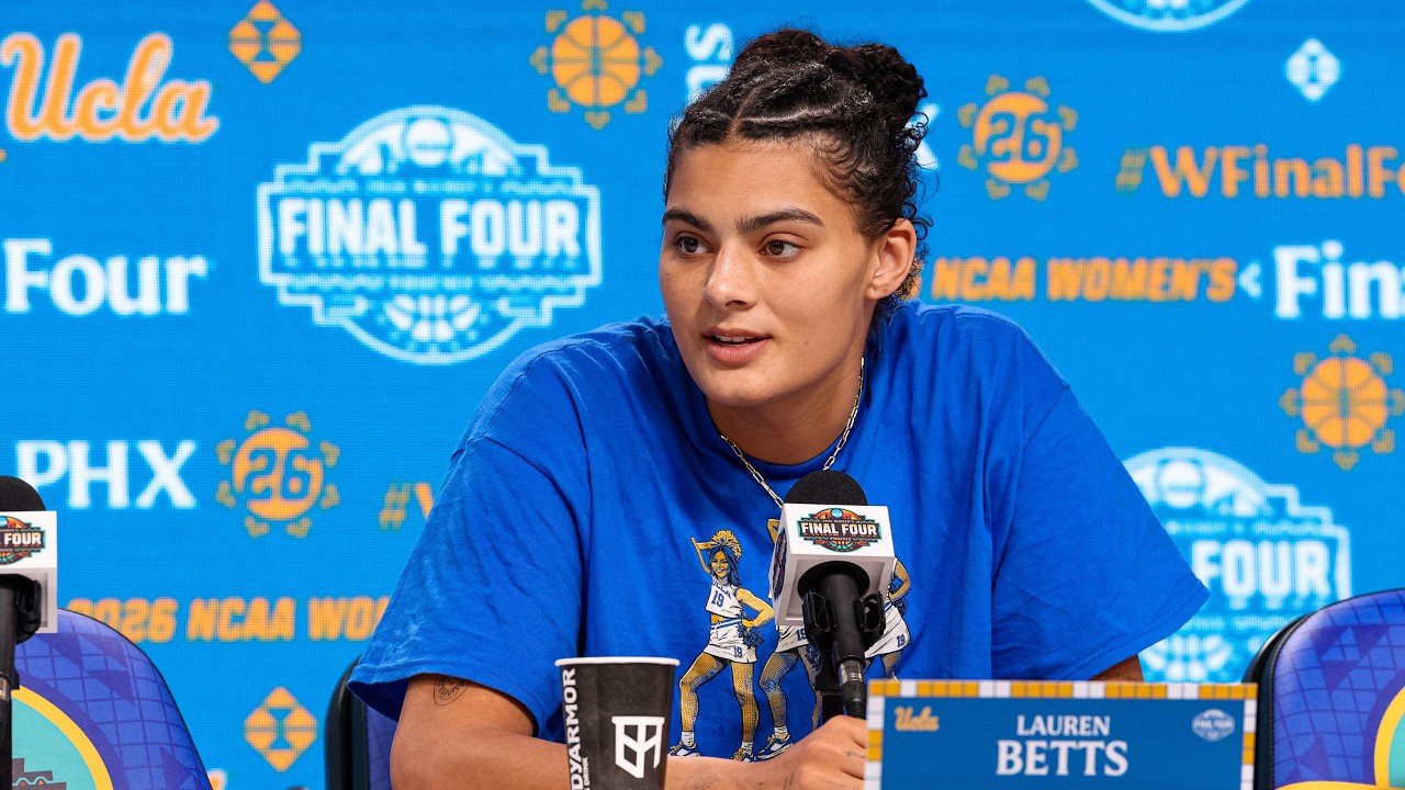 Basketball player Lauren Betts speaks at a press conference microphone with a blue NCAA Final Four backdrop behind her.