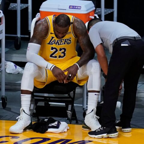 Basketball player in a Lakers yellow uniform sits on a bench, trainer leaning in as they inspect the ankle area on the floor.