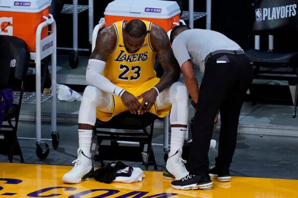 Basketball player in a Lakers yellow uniform sits on a bench, trainer leaning in as they inspect the ankle area on the floor.
