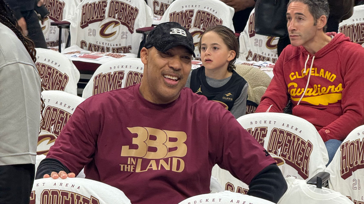 Smiling man in a maroon shirt sits in arena seats at a home opener, with a young girl behind him and another spectator nearby.
