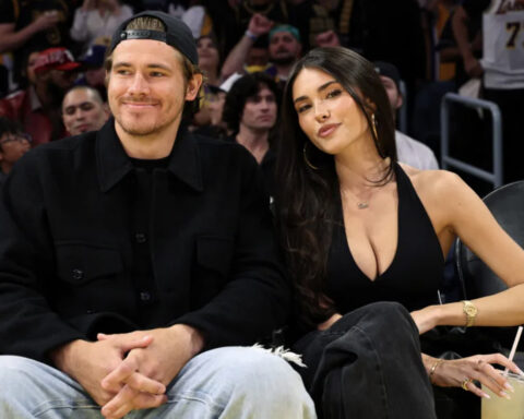 A man wearing a black jacket and cap sits beside a woman in a black sleeveless top at a sports arena, with a crowd in the background.",