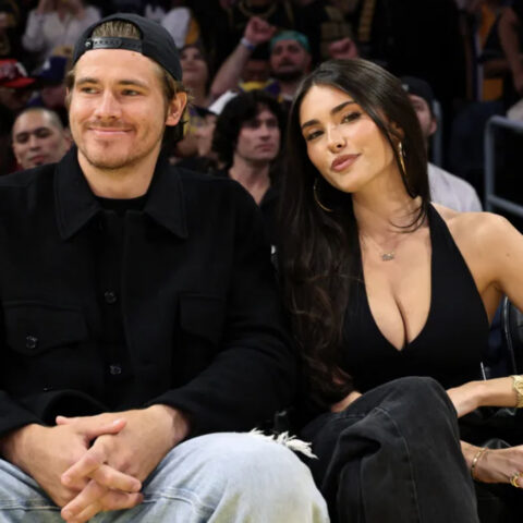 A man wearing a black jacket and cap sits beside a woman in a black sleeveless top at a sports arena, with a crowd in the background.",