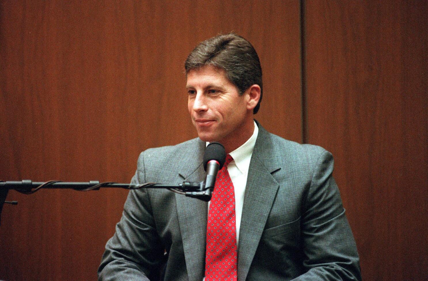 Man in a gray suit and red tie speaks into a microphone in a wood-paneled room.
