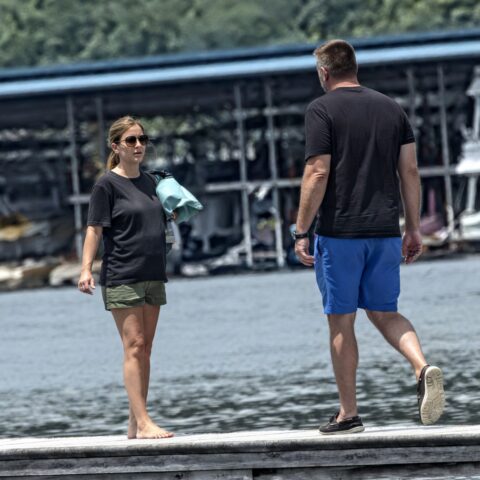 Two people stand on a weathered dock by a marina, the woman wearing sunglasses and carrying a light blue bag, while a man in blue shorts approaches from the right.