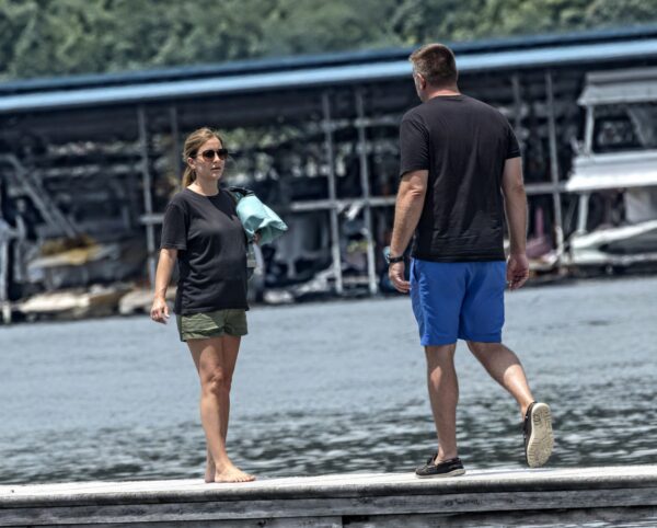 Two people stand on a weathered dock by a marina, the woman wearing sunglasses and carrying a light blue bag, while a man in blue shorts approaches from the right.