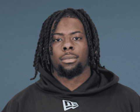 Portrait of a Black man with dreadlocks wearing a black hoodie against a blue-gray background, facing the camera with a neutral expression.