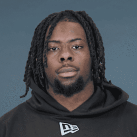 Portrait of a Black man with dreadlocks wearing a black hoodie against a blue-gray background, facing the camera with a neutral expression.