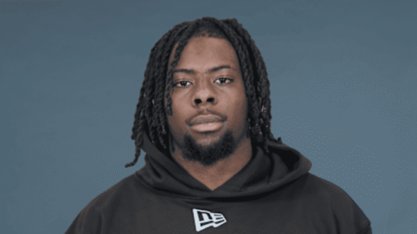 Portrait of a Black man with dreadlocks wearing a black hoodie against a blue-gray background, facing the camera with a neutral expression.
