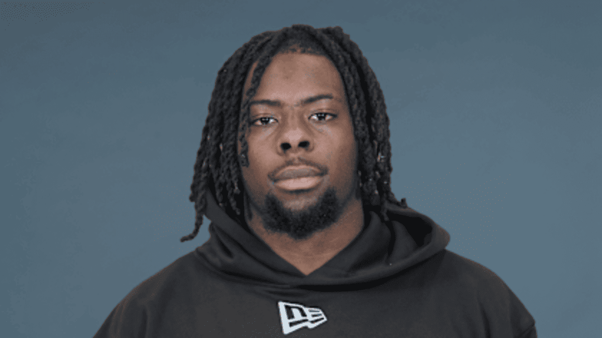 Portrait of a Black man with dreadlocks wearing a black hoodie against a blue-gray background, facing the camera with a neutral expression.