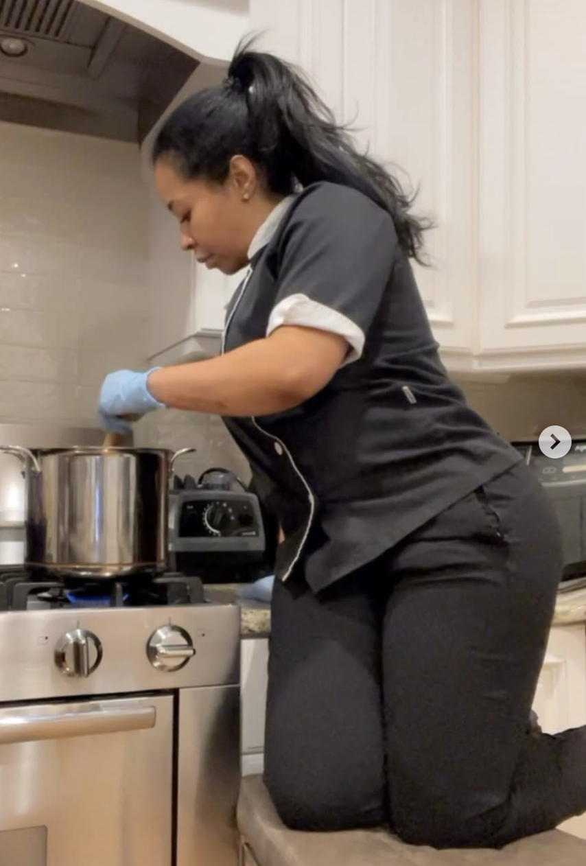 Woman in a black uniform kneels at a kitchen stove, stirring a large pot on a gas burner with a wooden spoon.