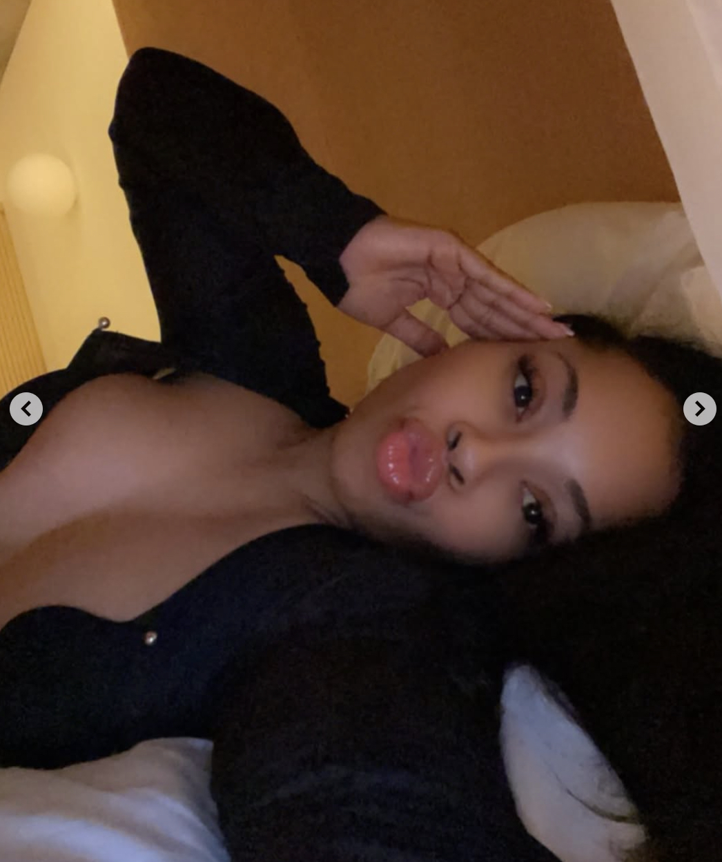 Woman lying on a bed, wearing a black top, lips puckered in a kissy pose with her hand by her head in dim lighting.