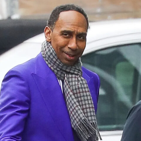 Man in a bright purple blazer and a plaid scarf smiles while standing near a white car outdoors on a city street.