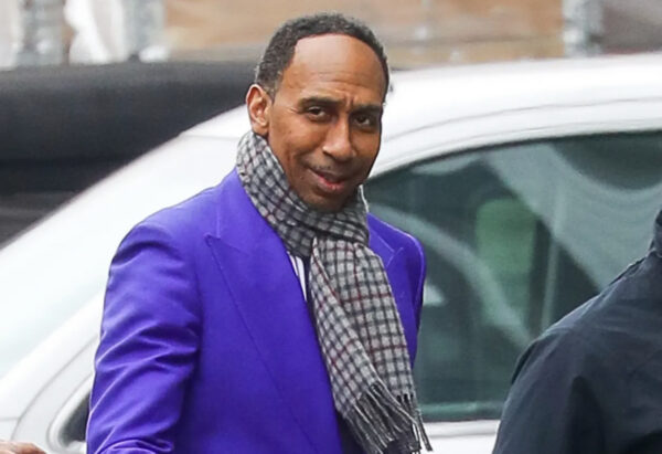 Man in a bright purple blazer and a plaid scarf smiles while standing near a white car outdoors on a city street.