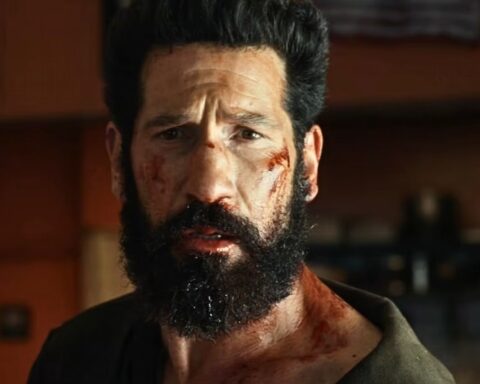Bearded man with blood on his face and neck, looking distressed in a dim indoor setting.
