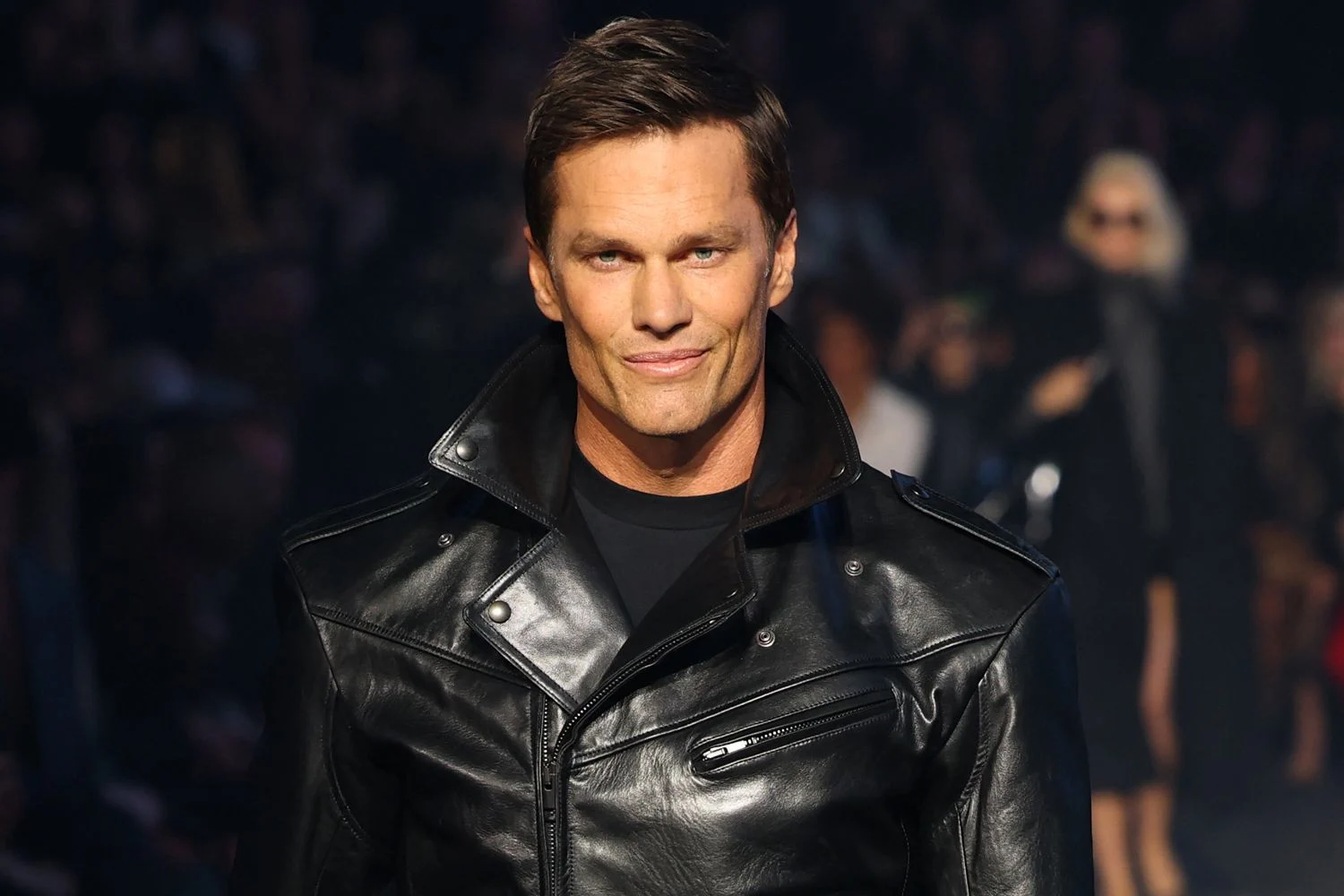 Male fashion model on a runway wearing a black leather jacket, posing with a slight smile as a dark audience backdrop is visible.
