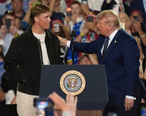 Two men on a stage with a lectern bearing the presidential seal; one in a black jacket shakes hands with a man in a blue suit as the crowd cheers.