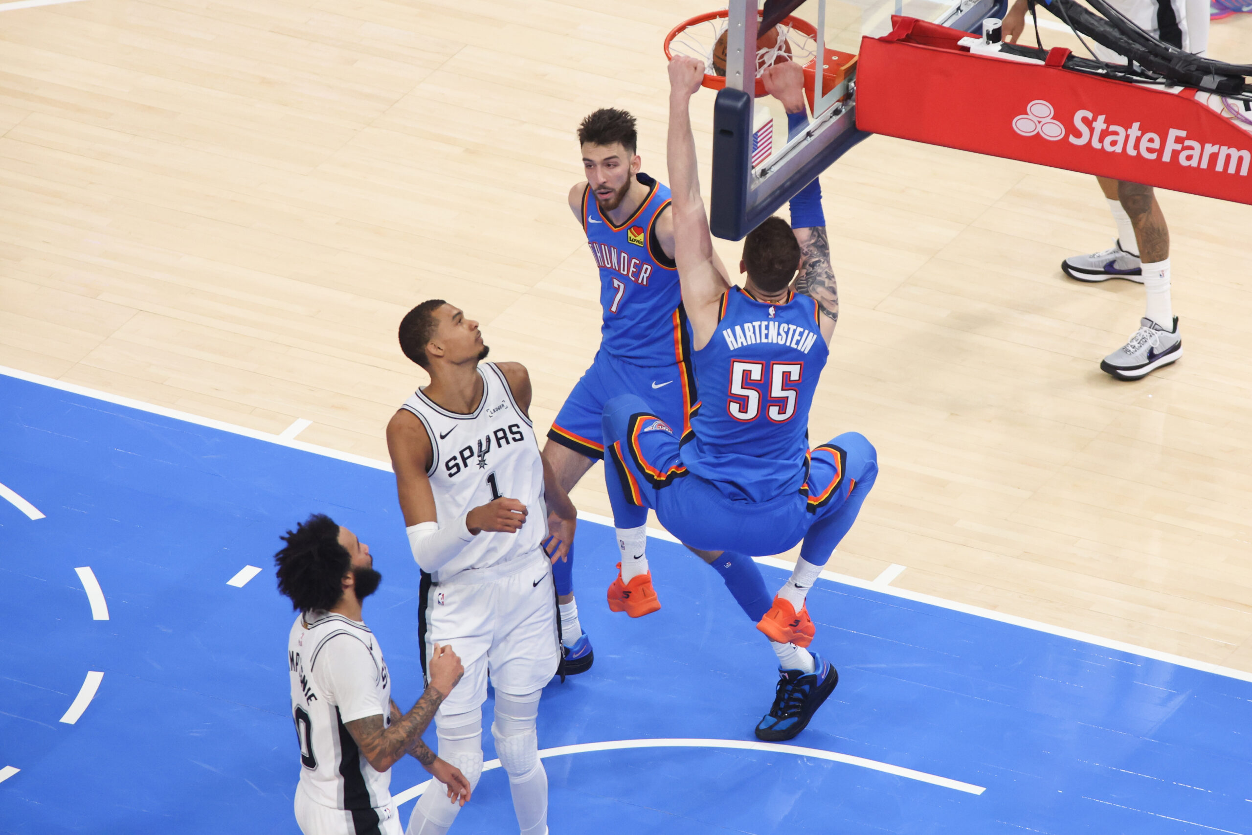 Basketball players in white Spurs uniforms guard the rim as two opponents in blue Thunder jerseys jump for a dunk near the hoop, with a State Farm padded backboard in view.