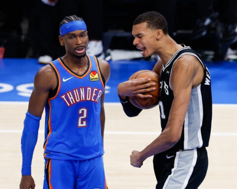 Two basketball players on the court during a game; one in a blue Thunder jersey with headband, the other in a black-and-white uniform holding the ball and talking.