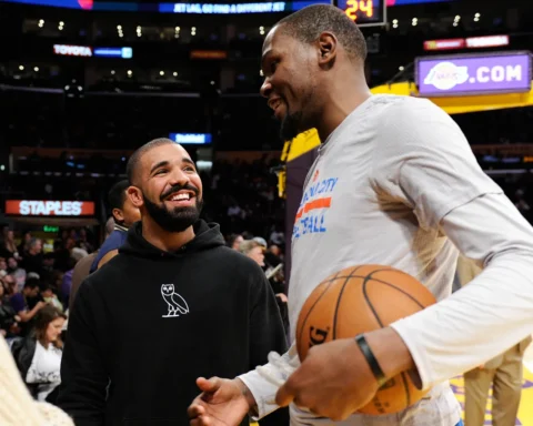 Two men on a basketball court share a smile: one in a white shirt holding a ball, the other in a black hoodie looking up at him. Fans backdrop in arena.