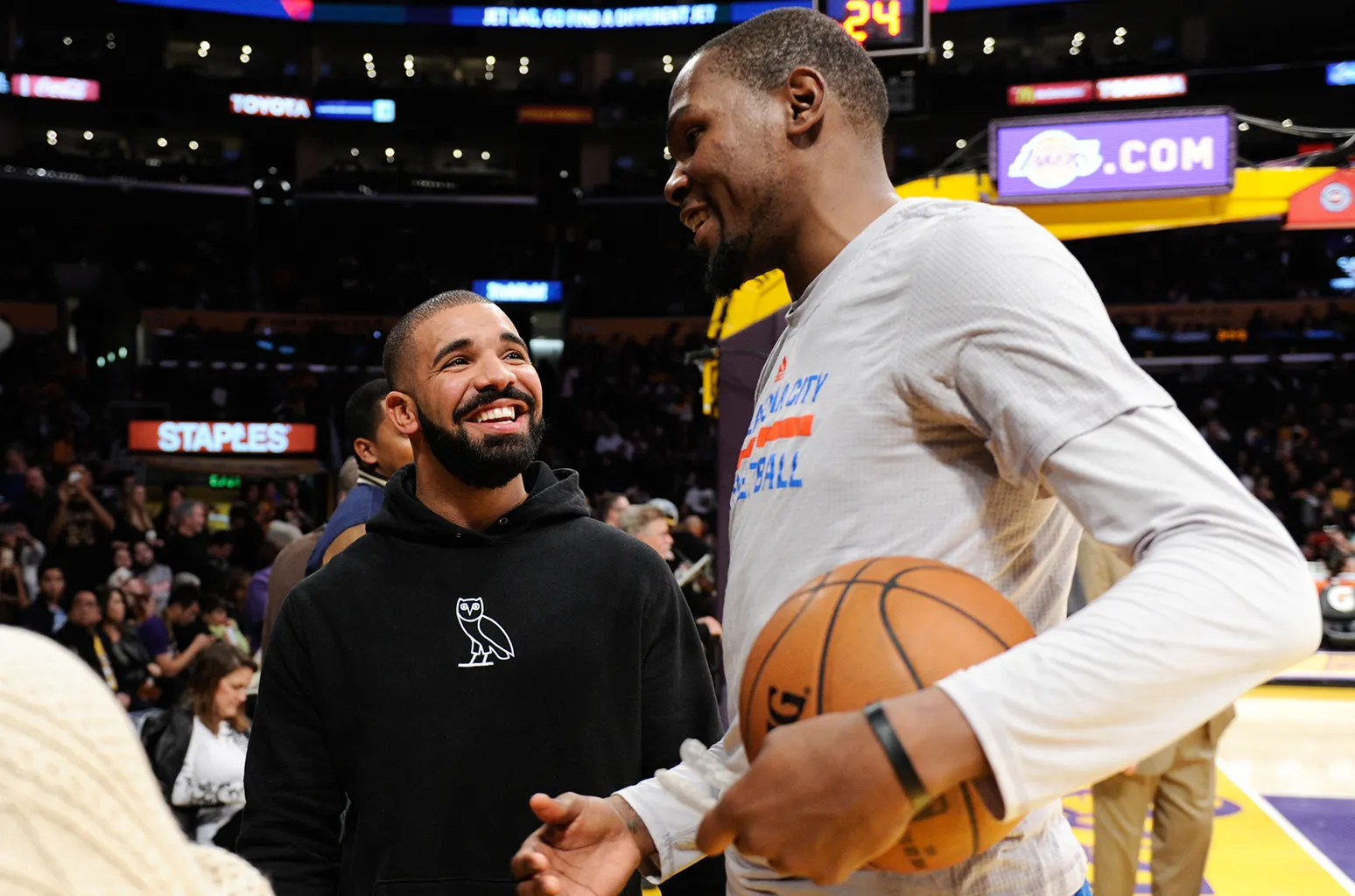 Two men on a basketball court share a smile: one in a white shirt holding a ball, the other in a black hoodie looking up at him. Fans backdrop in arena.