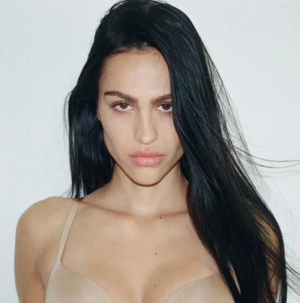 Portrait of a woman with long dark hair wearing a beige bra, looking at the camera against a light background.