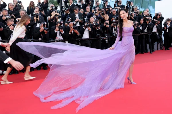 Woman in a lavender gown with a long flowing train posing on the red carpet as numerous photographers photograph her.
