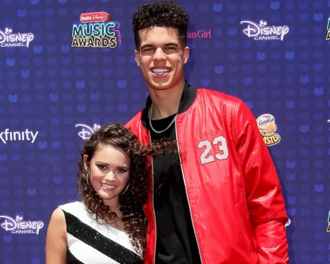 Two teens posing on a Disney Music Awards blue carpet; tall guy in a red jacket with the number 23 and a chain, a girl in a black-and-white striped dress next to him.