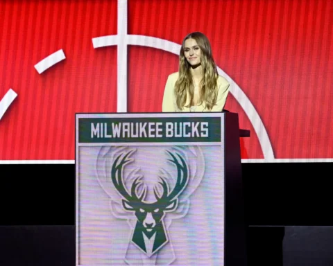 Woman with long blond hair in a light blazer stands at a podium with a Milwaukee Bucks logo on the front, red stage backdrop behind her.