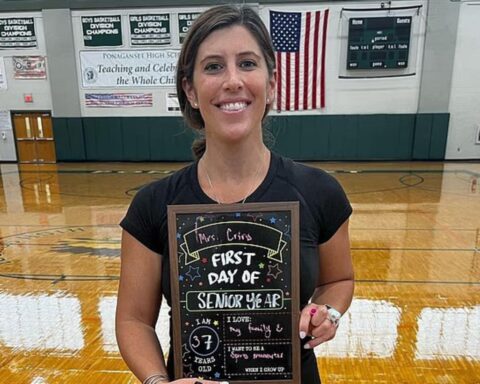 Woman in a gym holding a framed chalkboard that reads 'First Day of Senior Year' with a smile.