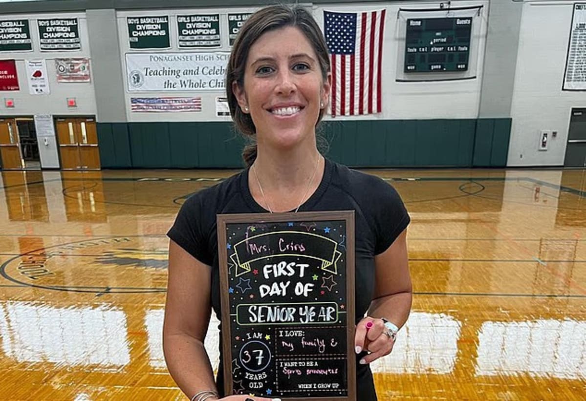 Woman in a gym holding a framed chalkboard that reads 'First Day of Senior Year' with a smile.