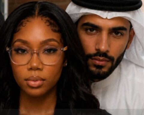 Close-up of a woman wearing clear-framed glasses with a man in a white kandura and headscarf behind her.
