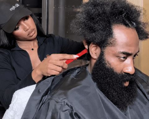 Stylist trimming a man's afro with a red comb while he wears a black cape in a salon.