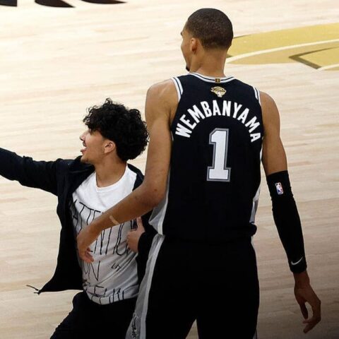 Two basketball players on a light wooden court; one in a Wembanyama #1 jersey stands while a friend in a jacket takes a selfie with a smartphone.