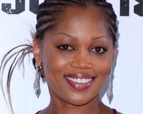 Close-up of a smiling woman with braided hair, diamond earrings, and glossy lipstick against a light background.