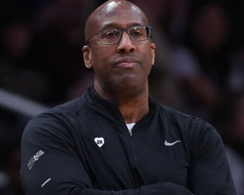 Man with short hair and glasses, wearing a black zip-up athletic jacket with a 'DM' pin, arms crossed and contemplative look in a arena.