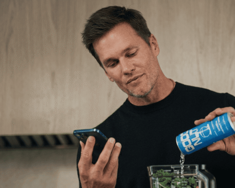 Man in a black sweater pours from a blue can into a blender while looking at his phone in a kitchen setting.