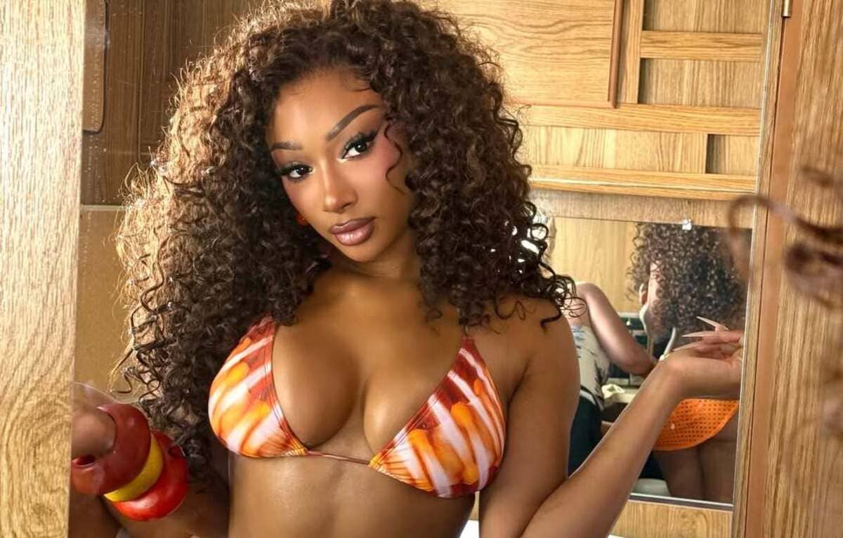 Woman with long curly hair wearing an orange-white patterned bikini top, posing in a wooden room with a mirror behind her.